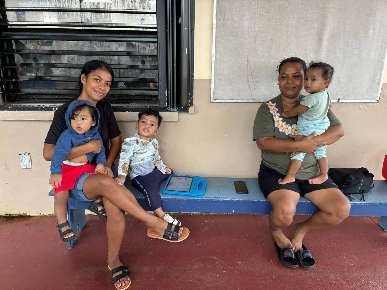 Two women sit on a wooden bench holding small children.