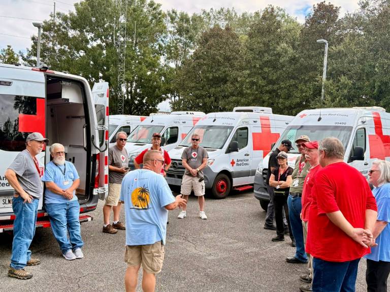 A group of people, many in Red Cross shirts, stand in front of Red Cross vehicles, listening to a man brief them on the disaster situation.