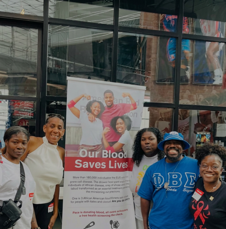Five adults smile in front of an American Red Cross banner reading "Our Blood Saves Lives" outside a building with large glass windows.