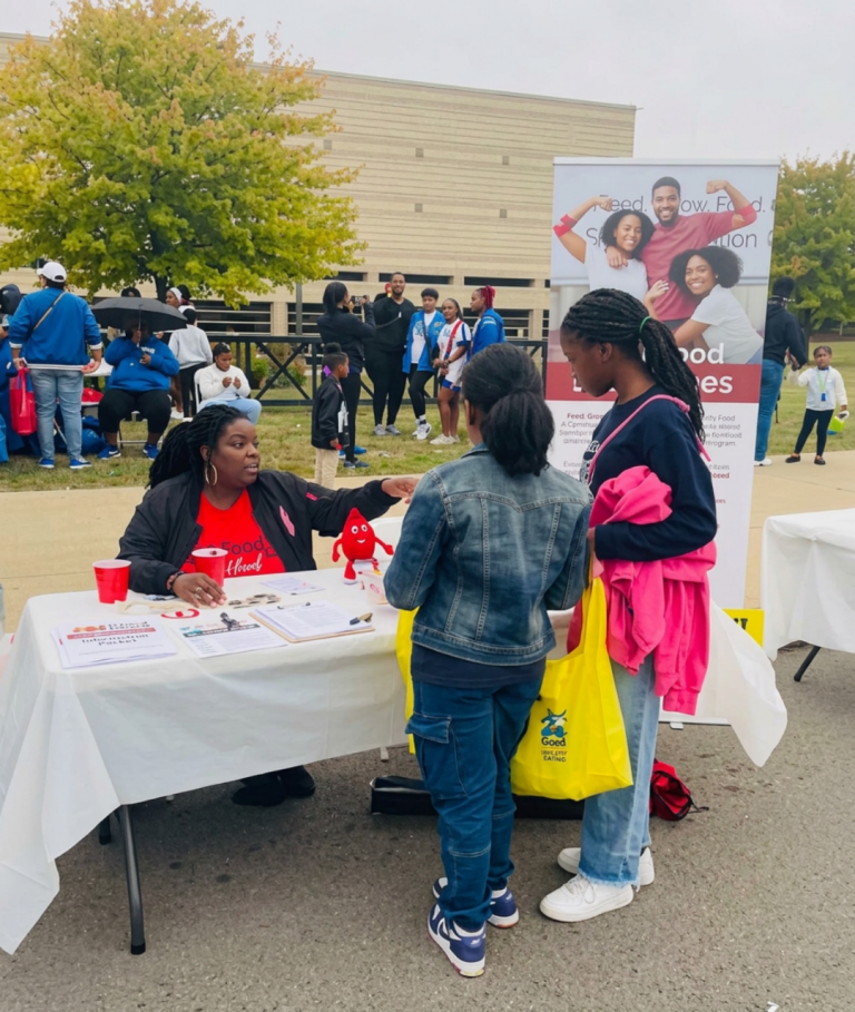 DeAndra Smith, seated at a white-draped recruitment table with printed materials and a red blood drop mascot, talks with two young visitors carrying tote bags at an outdoor event.