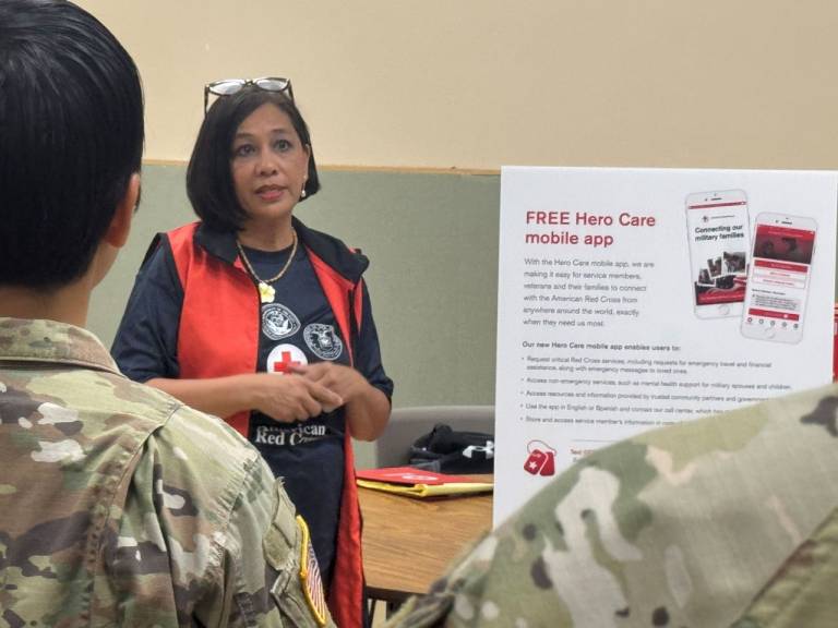 Woman in Red Cross vest stands in front of people in military uniforms.