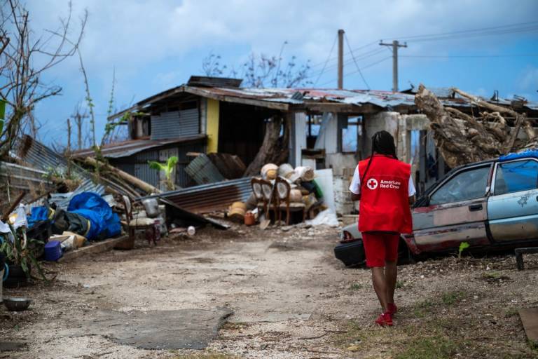 Woman in Red Cross vest walks along debris strewn street.