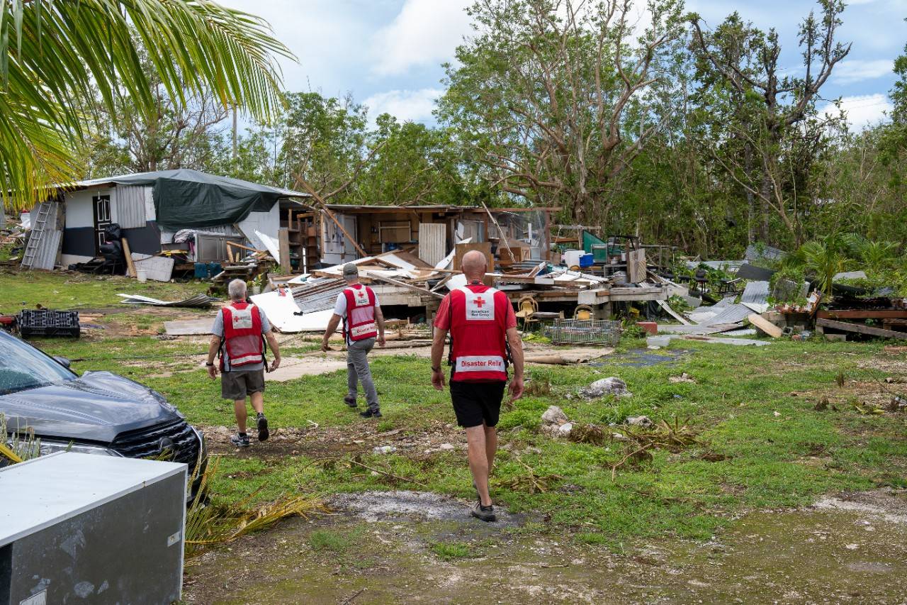 Three men in Red Cross attire walk through storm debris.