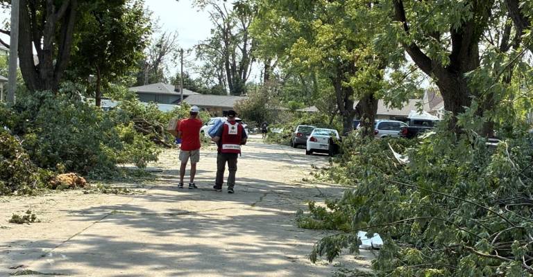 Man in Red Cross vest and man in red shirt walk down a street surrounded by downed tree branches.