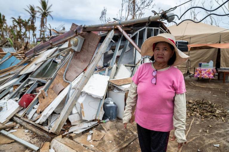 Woman in sun hat and pink shirt stands in front of destroyed home.