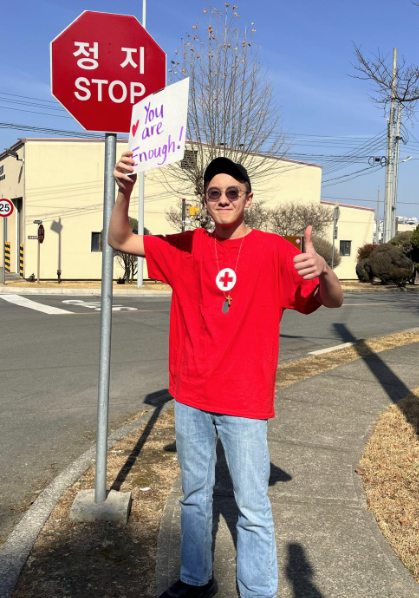 Un joven voluntario de la Cruz Roja Americana (Gyan) sostiene, por encima de la cabeza, un letrero que dice «You are Enough» y, con la otra mano, levanta el pulgar.