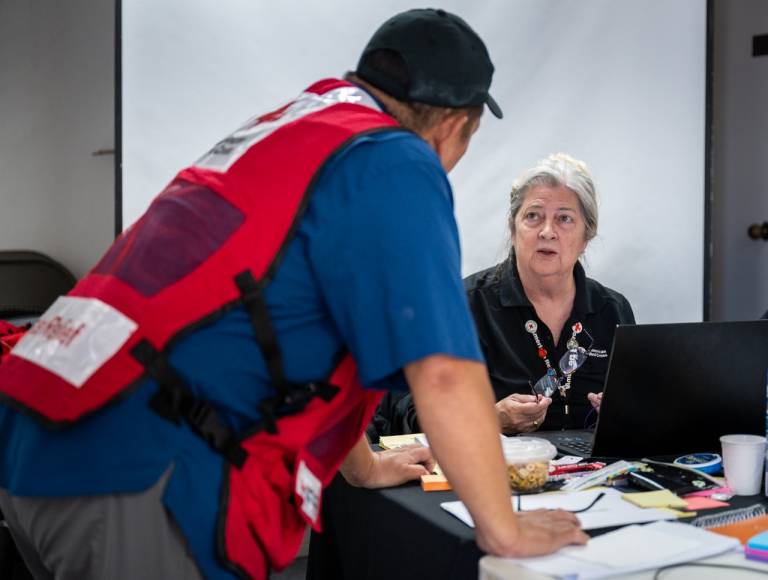 Woman in Red Cross vest sits at a desk talking to a man in a Red Cross vest