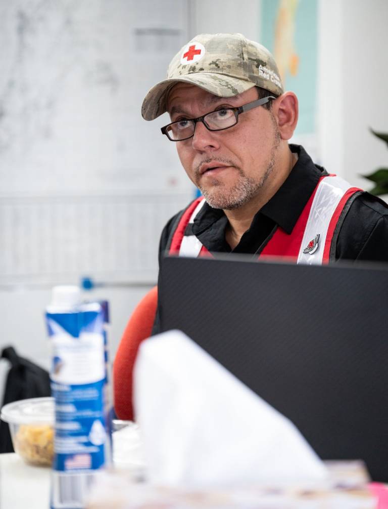Man in Red Cross baseball hat and vest sits behind a computer.