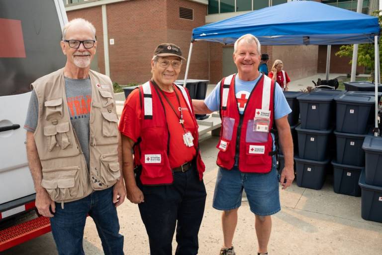 Three men in Red Cross vests stand outside a marked Red Cross vehicle.