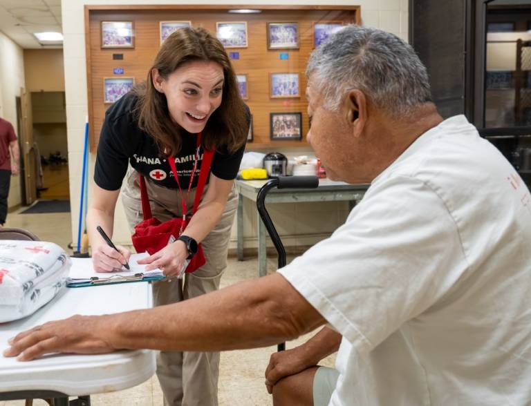 Woman in Red Cross shirt with clipboard and pen visits with a man in a Red Cross shelter