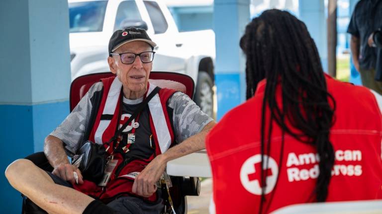 A man in a Red Cross vest and hat sits and talks with a volunteer in a Red Cross vest