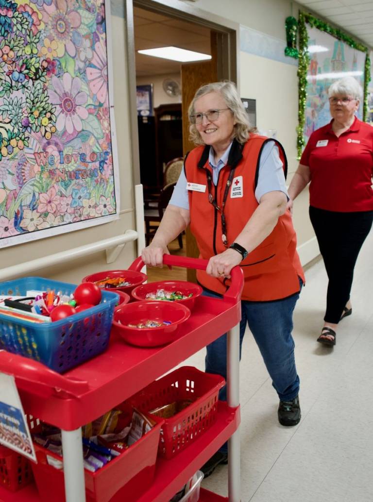 Woman in Red Cross vest pushes a cart down a hospital hallway filled with goodies for soldiers