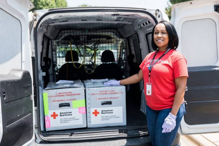 Woman in Red Cross tee shirt standing at van filled with blood boxes