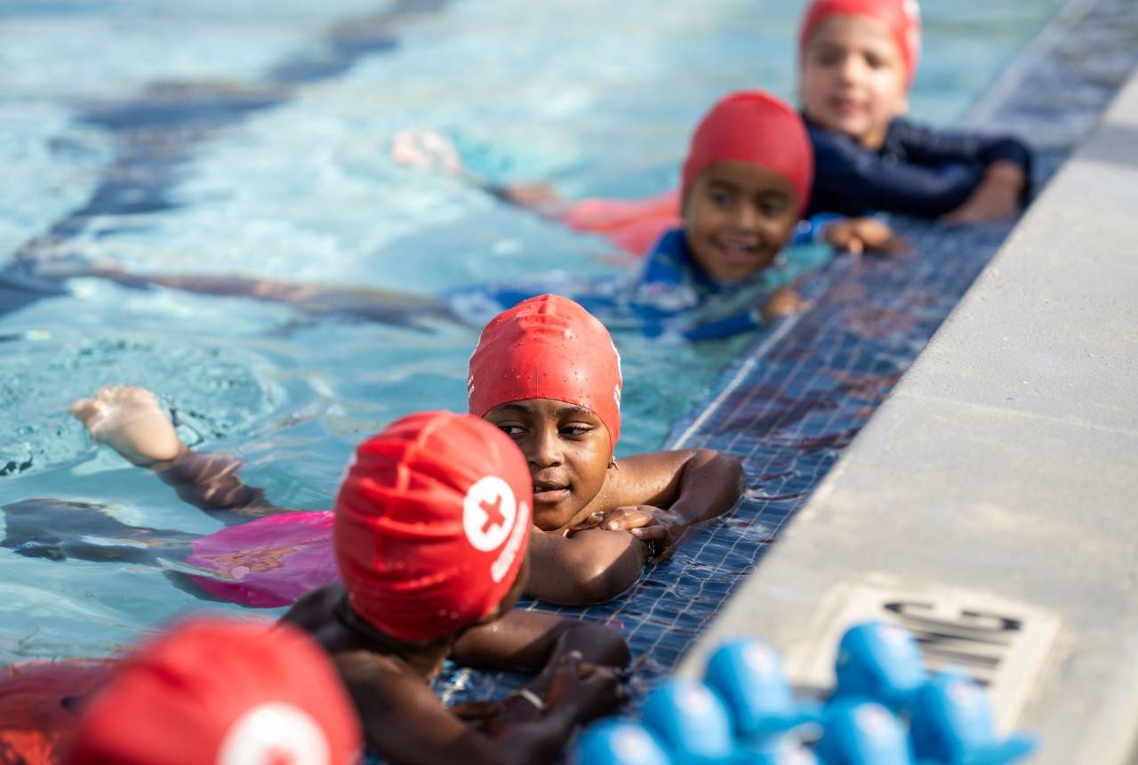 Children wearing red cross swim caps, hold onto the edge of a swimming pool during a swim lesson