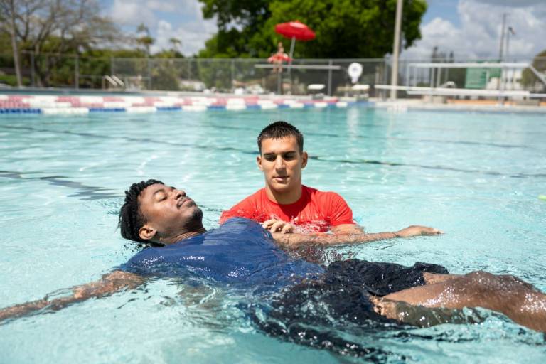 An American Red Cross instructor in a red shirt supports a participant as he practices floating on his back in an outdoor swimming pool during a swim lesson.
