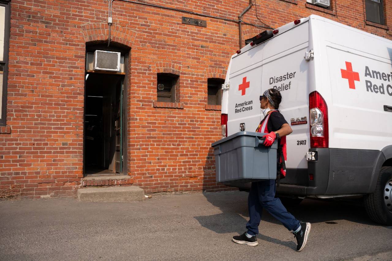 Person in Red Cross vest carries large tote toward a brick building.