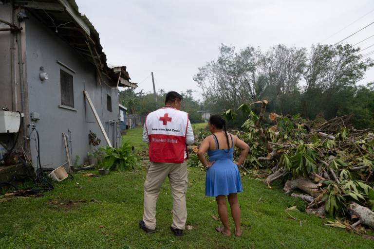 Man in Red Cross vest and woman in blue dress study her damaged home.