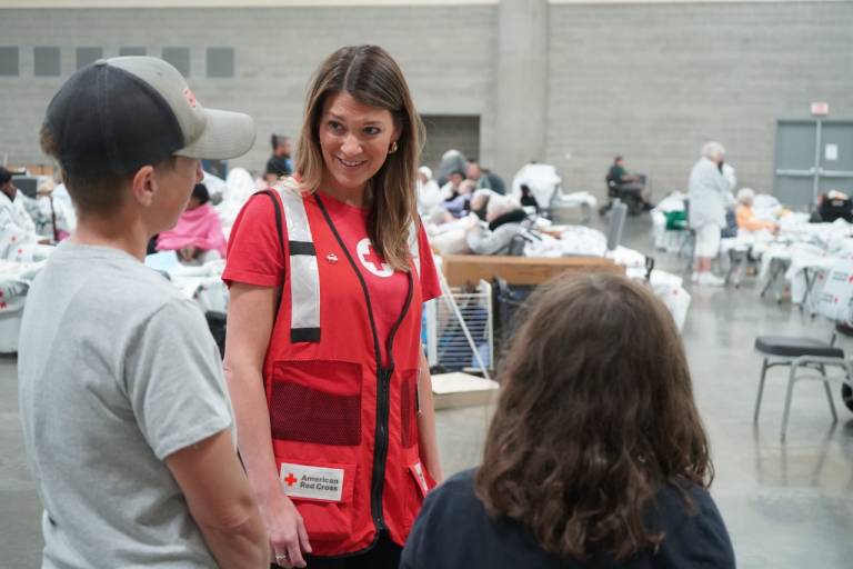 Woman in Red Cross vest talks with two people in a shelter.