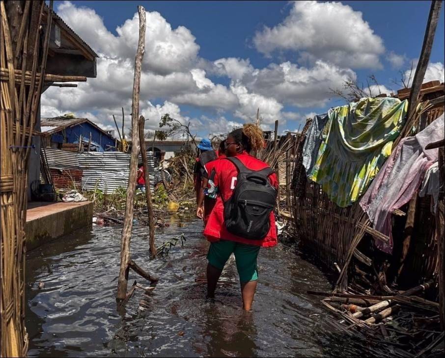 Alt text – people in Red Cross vests wade through water, surrounded by damaged buildings