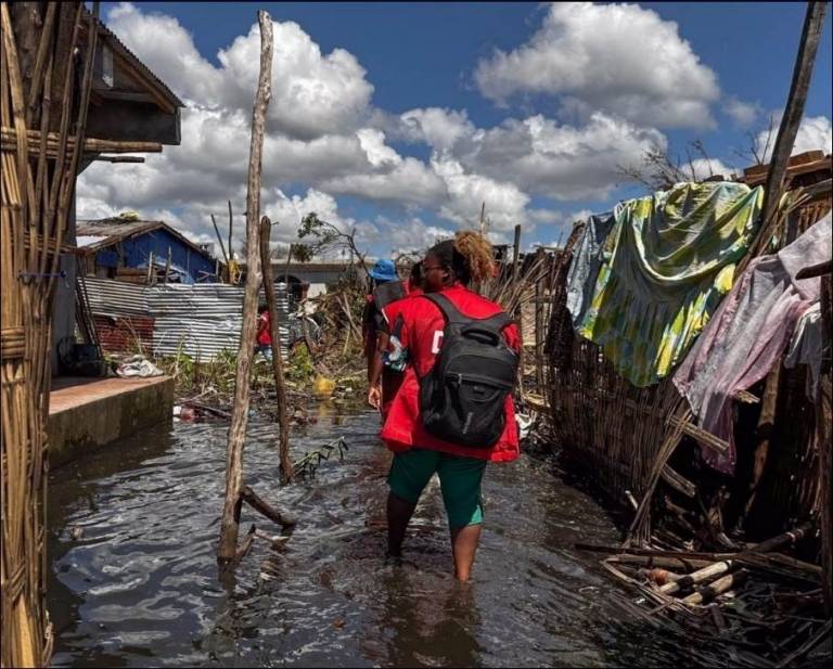people in Red Cross vests wade through water, surrounded by damaged buildings