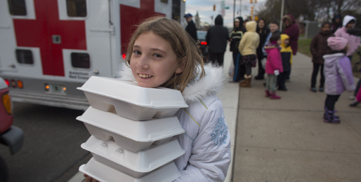 Young girl carrying food in foam packaging with people in background next to Red Cross vehicle