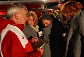 American Red Cross volunteer holding a clipboard and pen talking with 3 people with blankets wrapped around them.