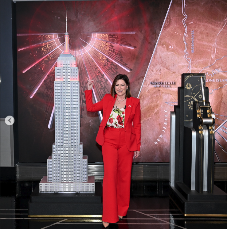 Chloe Melas pointing up toward the top of a large model of the Empire State building  lit up with red lights