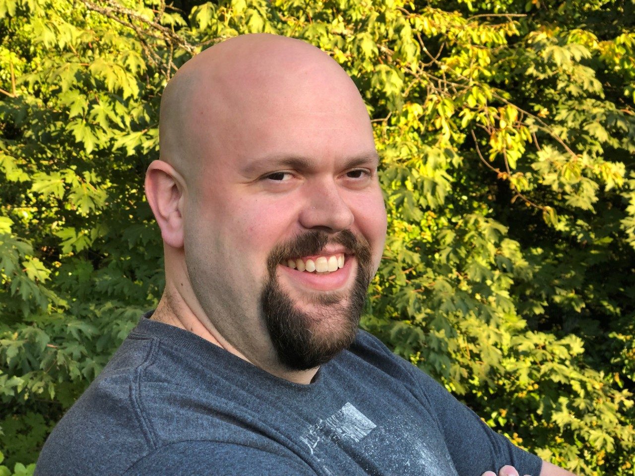 Smiling man with light skin and brown facial hair stands in front of a green space. 