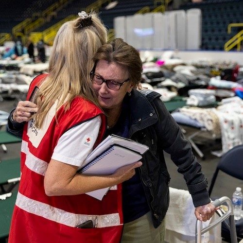 A blonde Red Cross volunteer is hugging an older woman in a shelter.