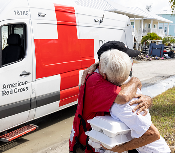 A Red Cross volunteer hugs a client in front of an emergency response vehicle.