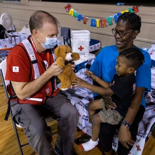 Valerie and her son are being entertained by a Red Cross volunteer with a teddy bear in a shelter.