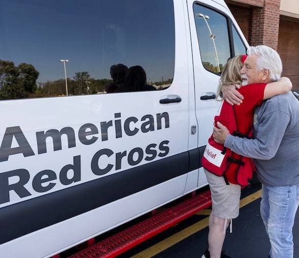 A Red Cross volunteer hands a client a warm meal in front of an emergency response vechicle showing the text "All American Red Cross dusaster assistance is free."