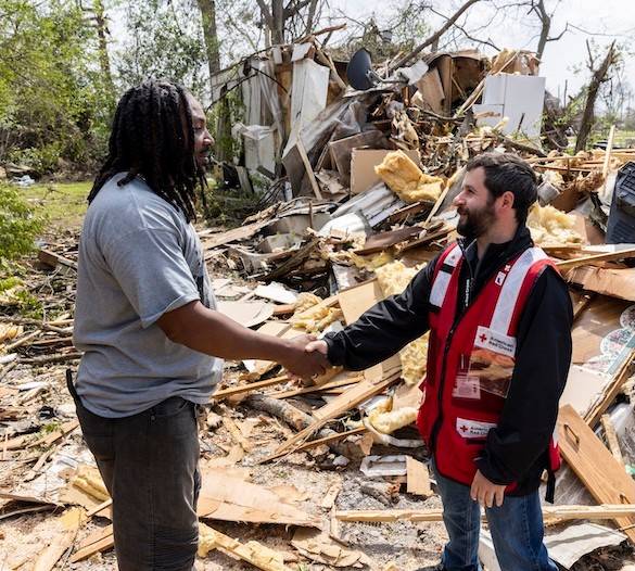 A Red Cross disaster assistance volunteer shakes hands with a client in front of a home destroyed by a tornado.