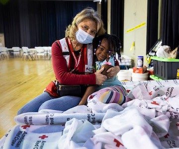 Masked volunteer in Red Cross shelter hugs a smiling young girl