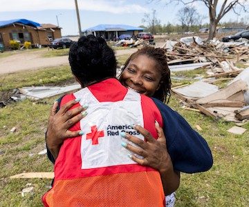 Volunteer and woman hug while standing outside amidst a neighborhood affected by a tornado