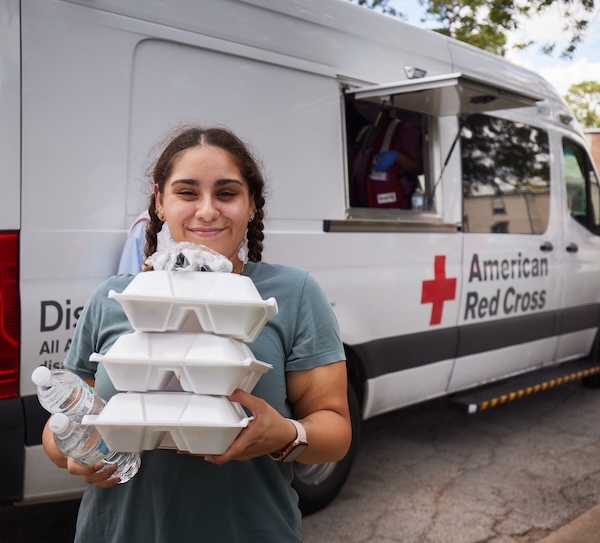 A young girl holds several food containers while standing in front of an American Red Cross emergency response vehicle. 