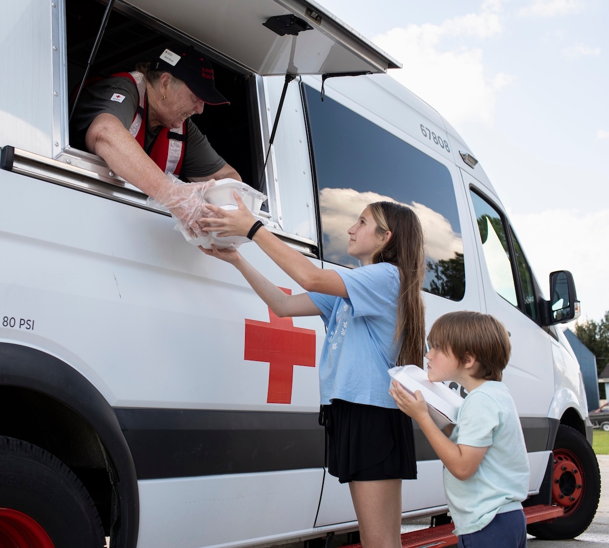 Two children receive meals from a Red Cross worker through a mobile relief vehicle window.