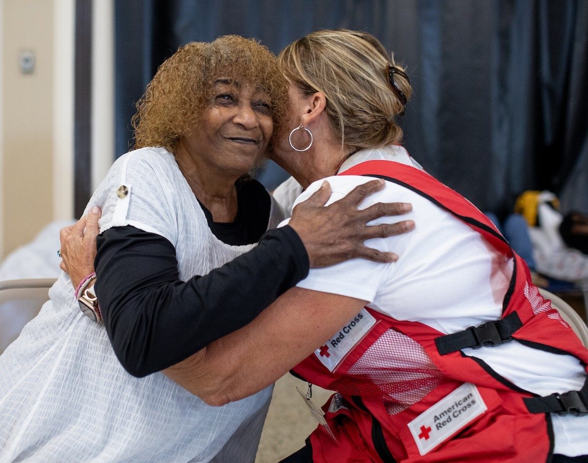 A Red Cross volunteer embraces an older individual in a community space.