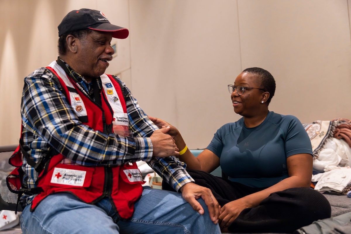 A Red Cross volunteer speaks with a seated individual in an indoor shelter setting.