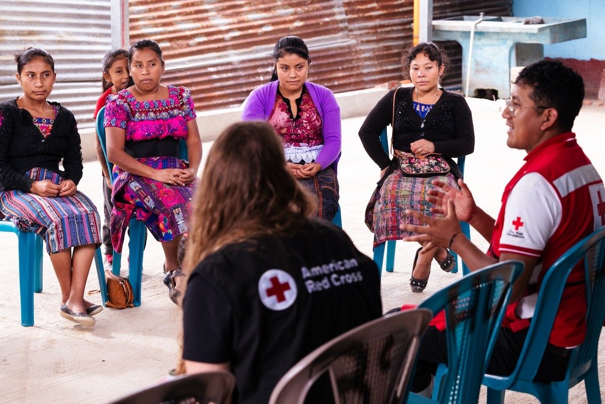 Red Cross workers lead a group discussion with seated participants in an open-air setting.