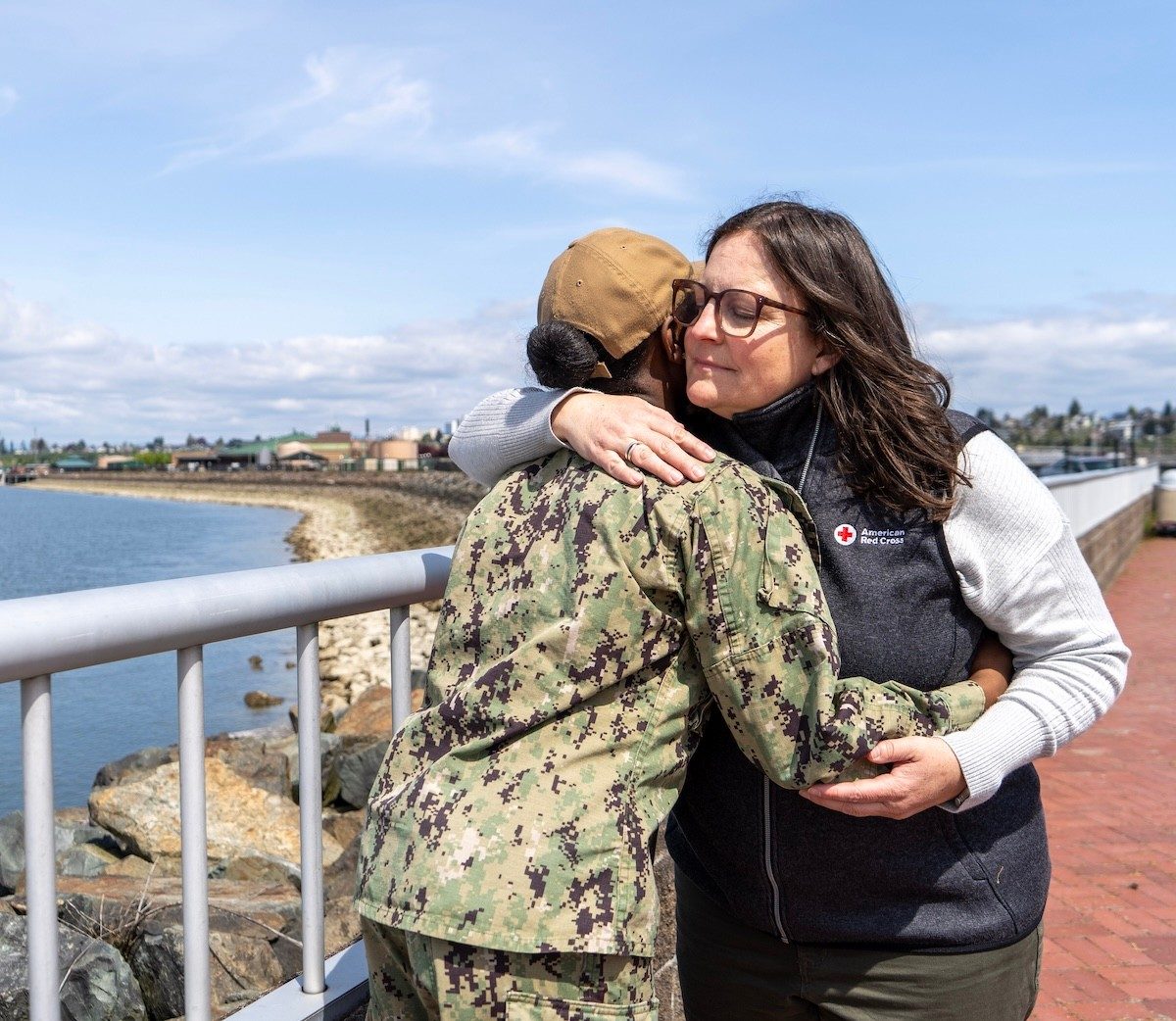 Two individuals embrace by a waterfront, one in a military-style uniform and the other in Red Cross outerwear.
