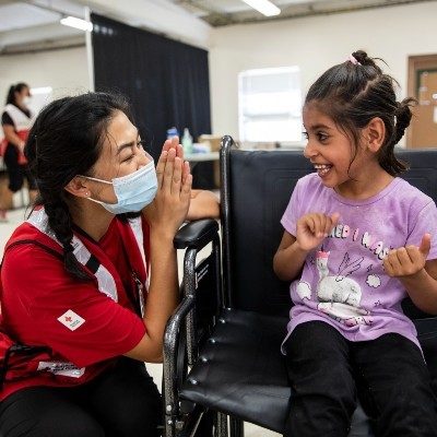 Red Cross volunteer wearing mask next to young girl in wheelchair who is smiling.