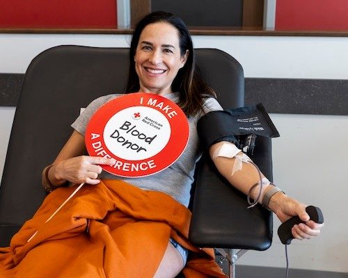 A blood donor with long, dark hair sits in a chair while in the process of donating blood. She holds a sign reading I Made a Difference - Blood Donor