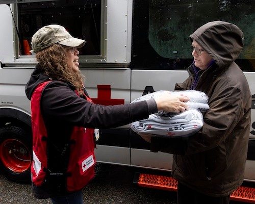Red Cross volunteer in red vest and tan hat stands next to an emergency response van handing blankets to a client