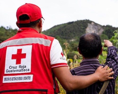 image of a back view of a volunteer wearing a Cruz Roja Guatemalteca vest as he and a client overlook rolling green hills.