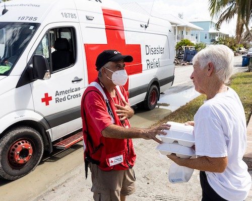 red cross volunteer standing next to a van handing food and supplies to someone