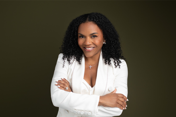 Ellen Rucker Sellers, a woman with natural, curly, dark brown hair, smiles in a white suit standing with her arms crossed in front of her.
