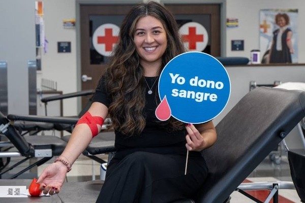 Marilyn Jiminez, a latina with long, dark hair, smiles while holding a sign reading yo dono sangre, and showing off her bandaged arm after donating blood.