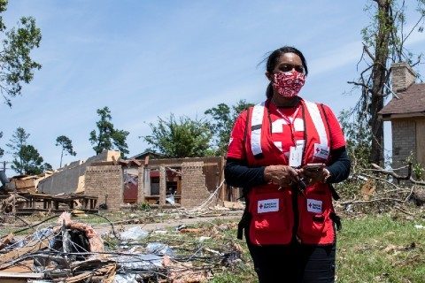 Red Cross volunteer wearing a mask and Red Cross vest standing by a house that is demolished.