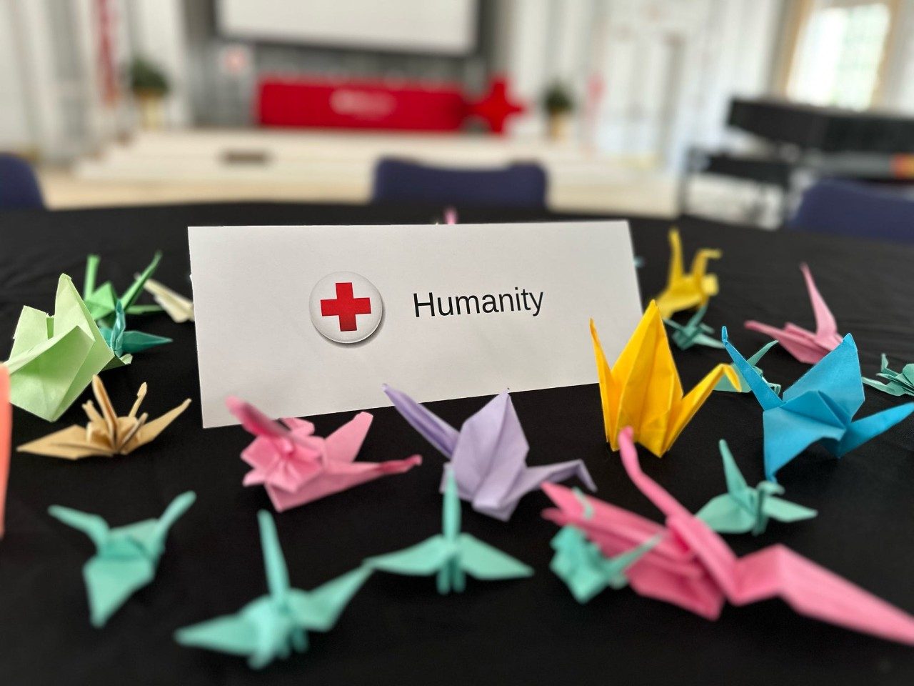 a small card with an American Red Cross symbol on it that says "Humanity", surrounded on a clothed table with tiny paper cranes